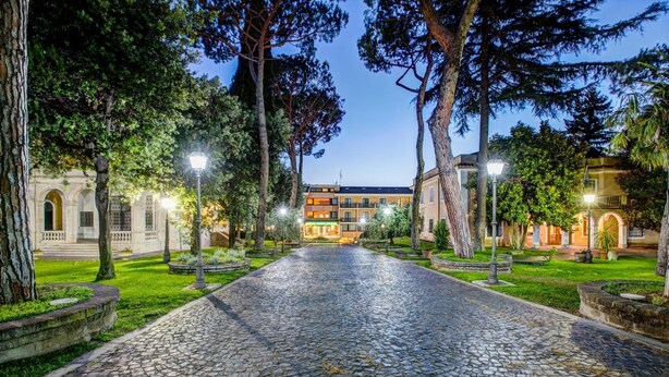 A cobblestone pathway leading to a building with a central courtyard, surrounded by trees and street lamps. - Appia Park Hotel (Rome)
