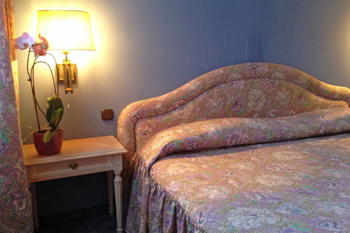 Un dormitorio con una colcha estampada, una mesita de noche con una lámpara y una orquídea en maceta, y una lámpara de pared.