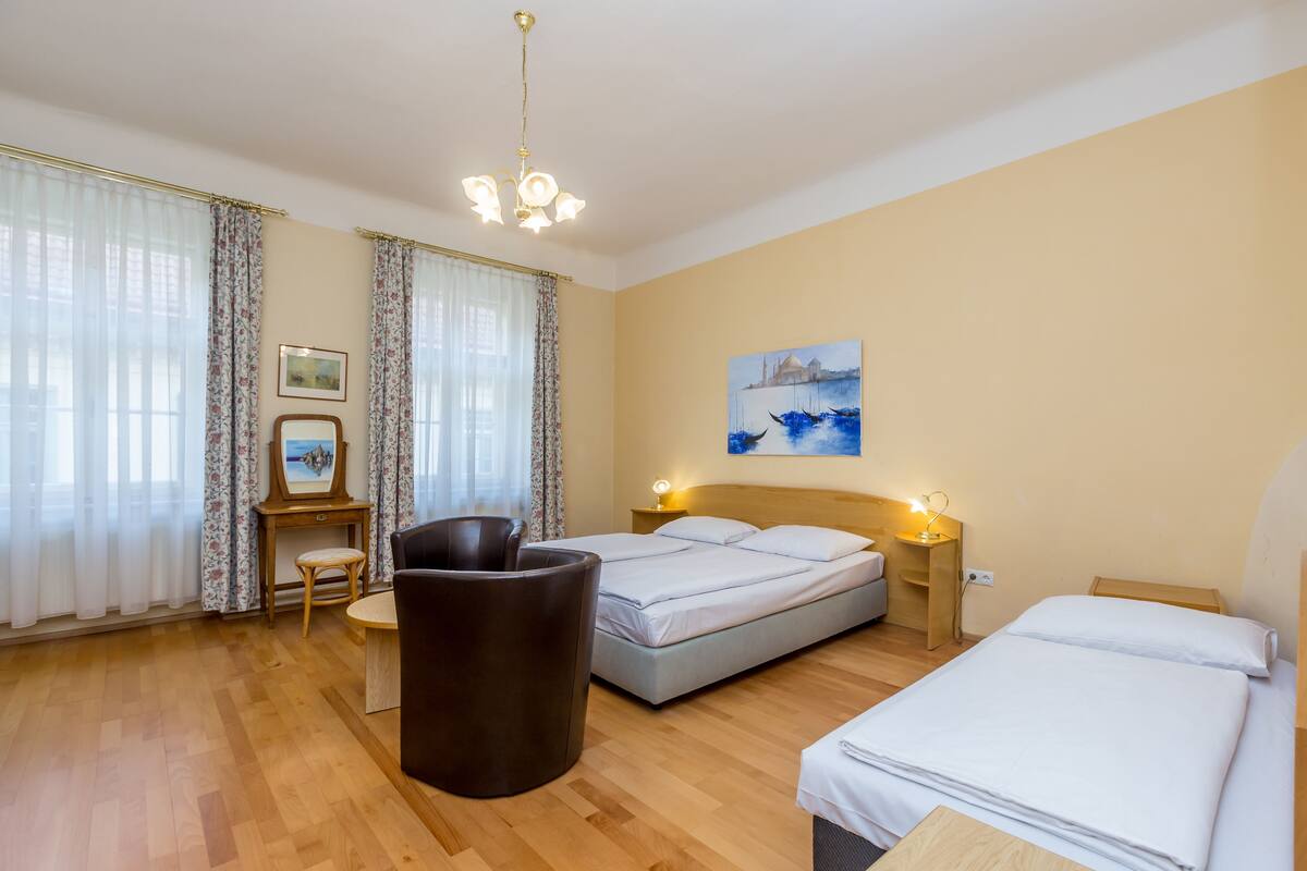 Une chambre d’hôtel avec un plancher en bois, deux lits, un bureau avec une chaise, une petite table, un tableau et un lustre.