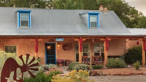 A rustic building with a metal roof, two blue-framed windows, a front door, and a porch with outdoor seating. - Casa Escondida Bed & Breakfast (Chimayo)