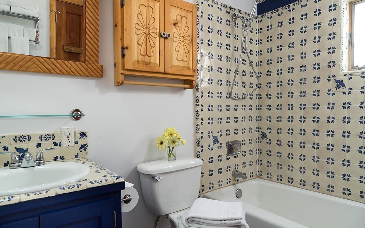 Un baño con lavamanos, inodoro y bañera blancos. Hay un mueble de madera con detalles florales sobre el inodoro y un espejo sobre el lavamanos. Las paredes están revestidas con un estampado floral.