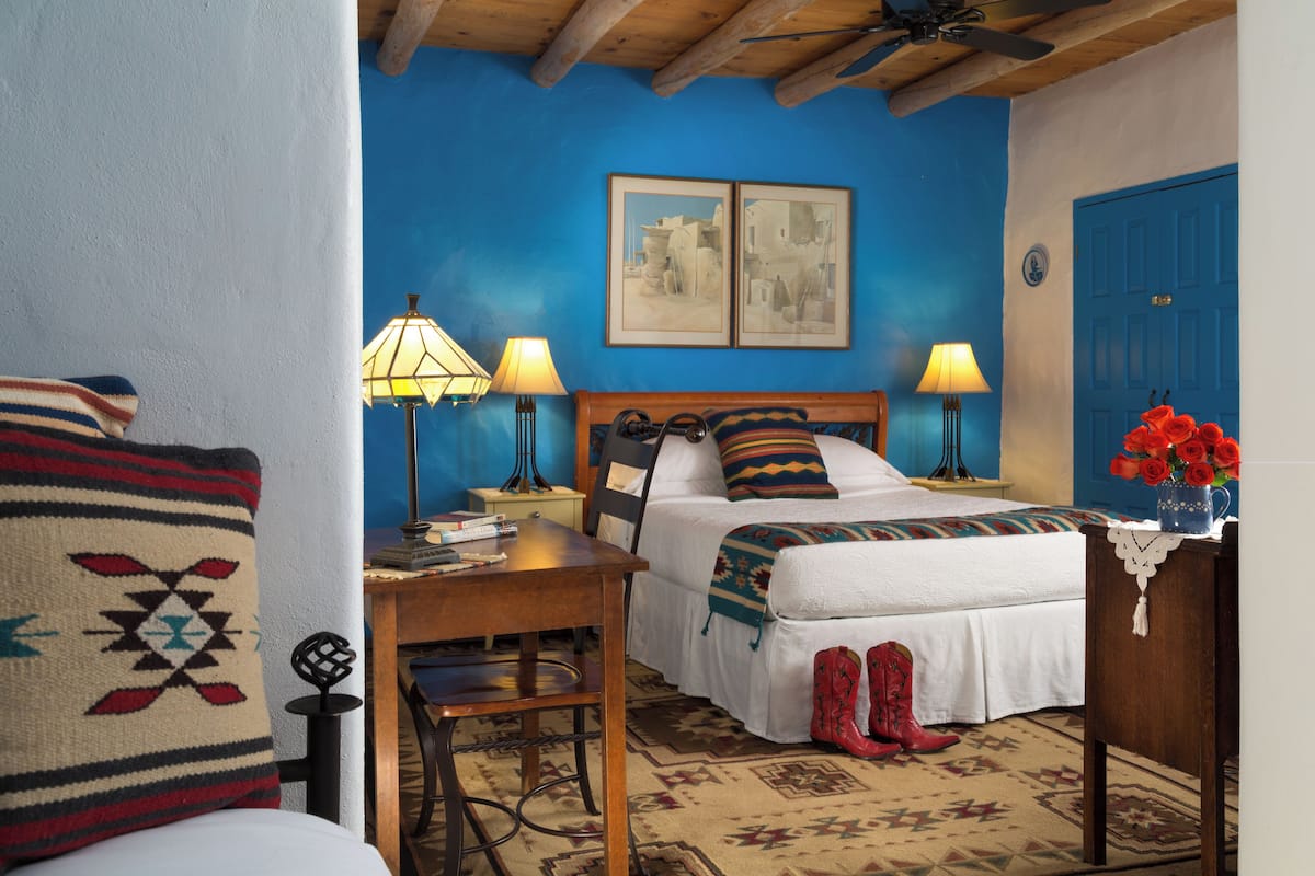 Une chambre à coucher avec un lit, deux lampes de chevet, une table de nuit en bois, un bureau avec une chaise, un tapis à motifs et une porte avec une poignée bleue.