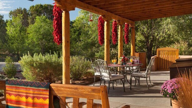 An outdoor patio with wooden furniture, a table set with a vase of flowers, and a wooden bench with a colorful blanket. - Casa Escondida Bed & Breakfast (Chimayo)