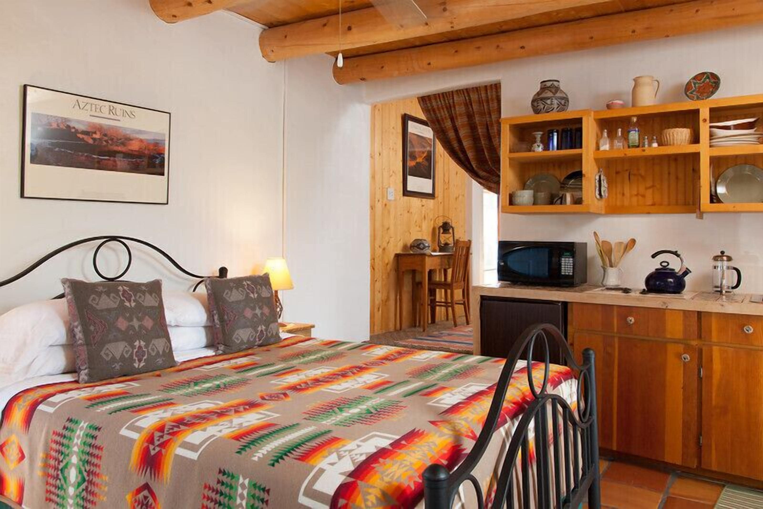A room with a bed, a wooden desk, a microwave, and shelves with kitchenware.