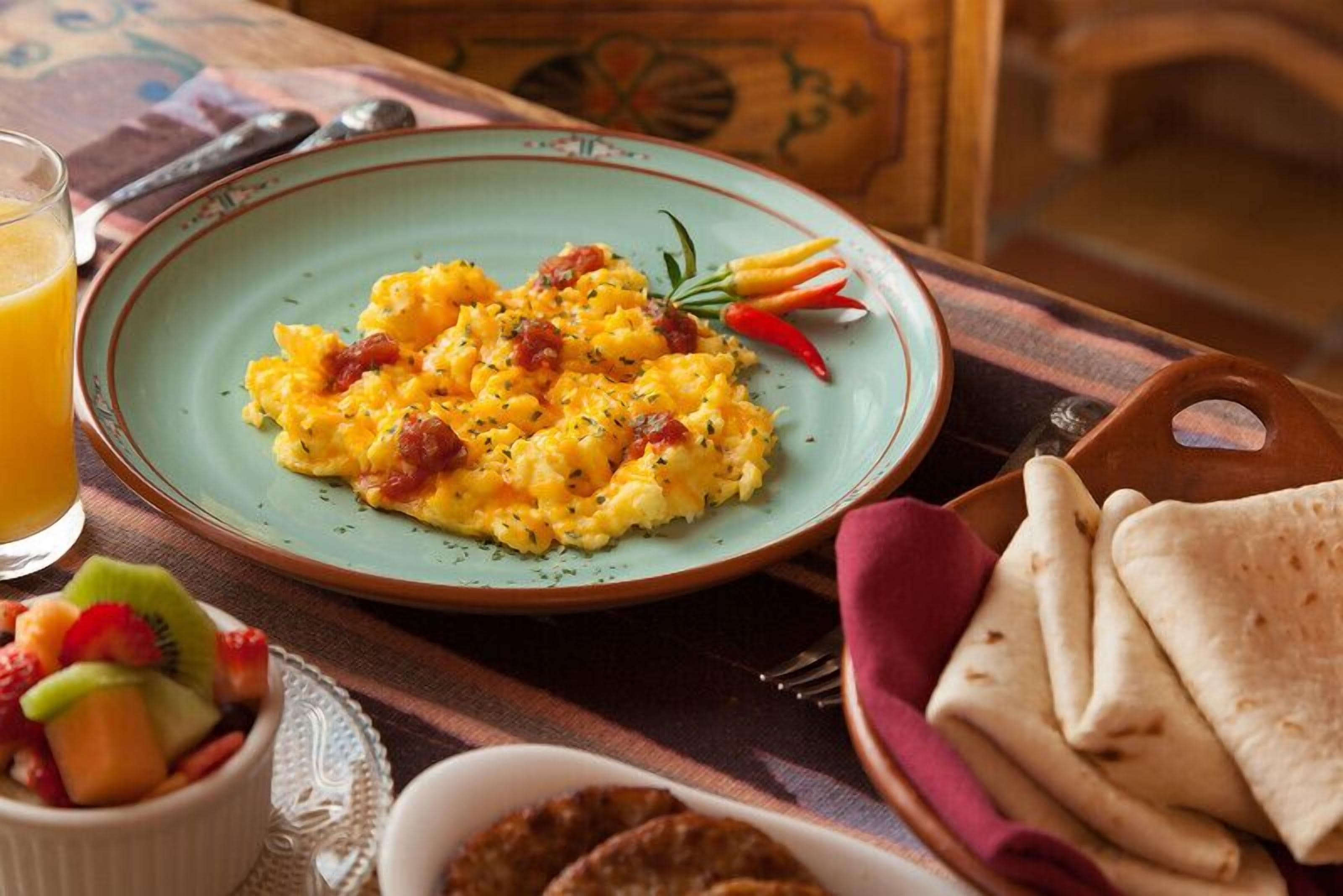 A breakfast plate with scrambled eggs, bacon, and herbs, accompanied by a glass of orange juice, a fruit bowl, and flatbread on a table.