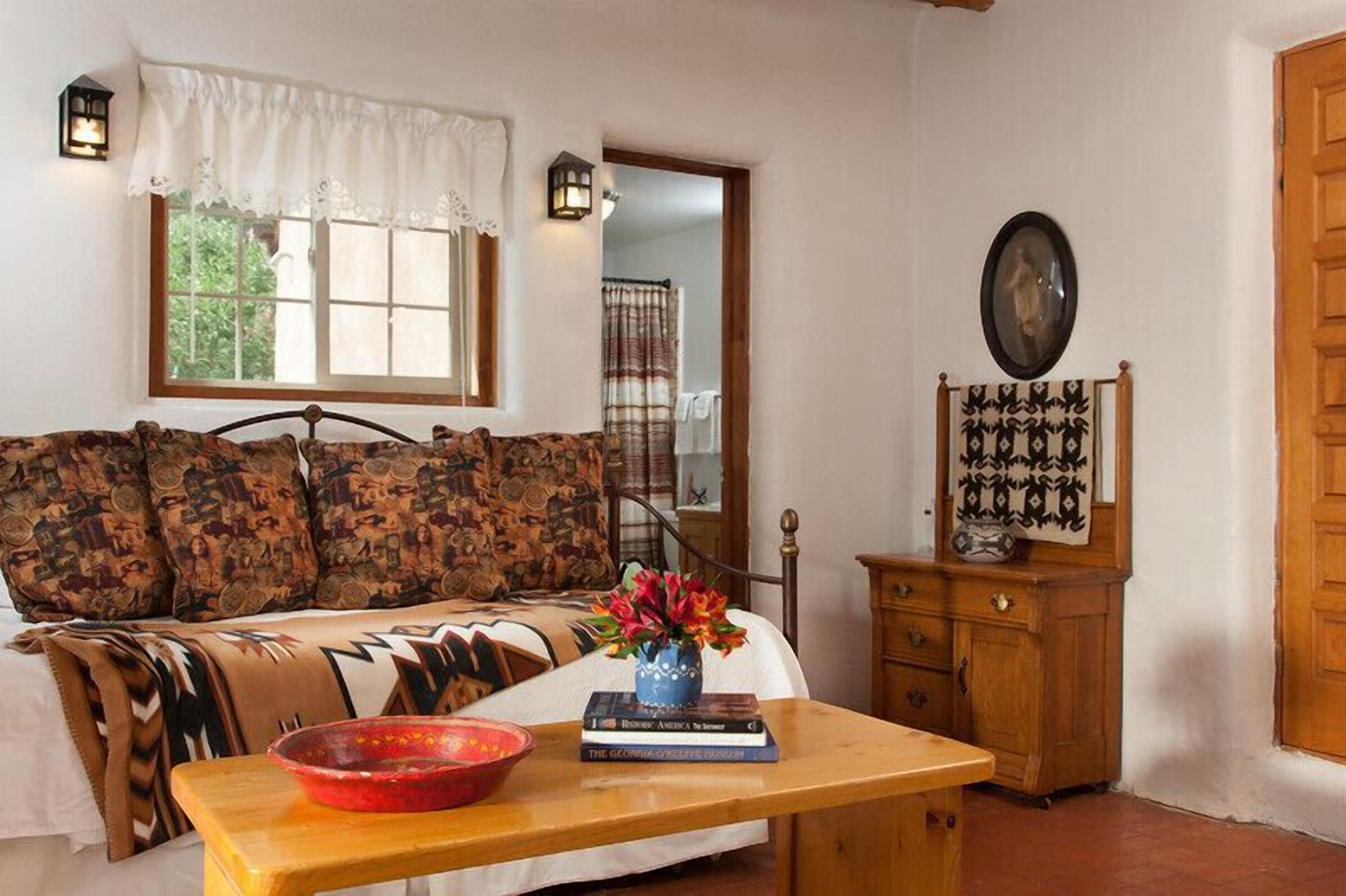 A cozy room with a bed, a wooden table with a bowl and books, a dresser, a window with curtains, and a mirror on the wall.