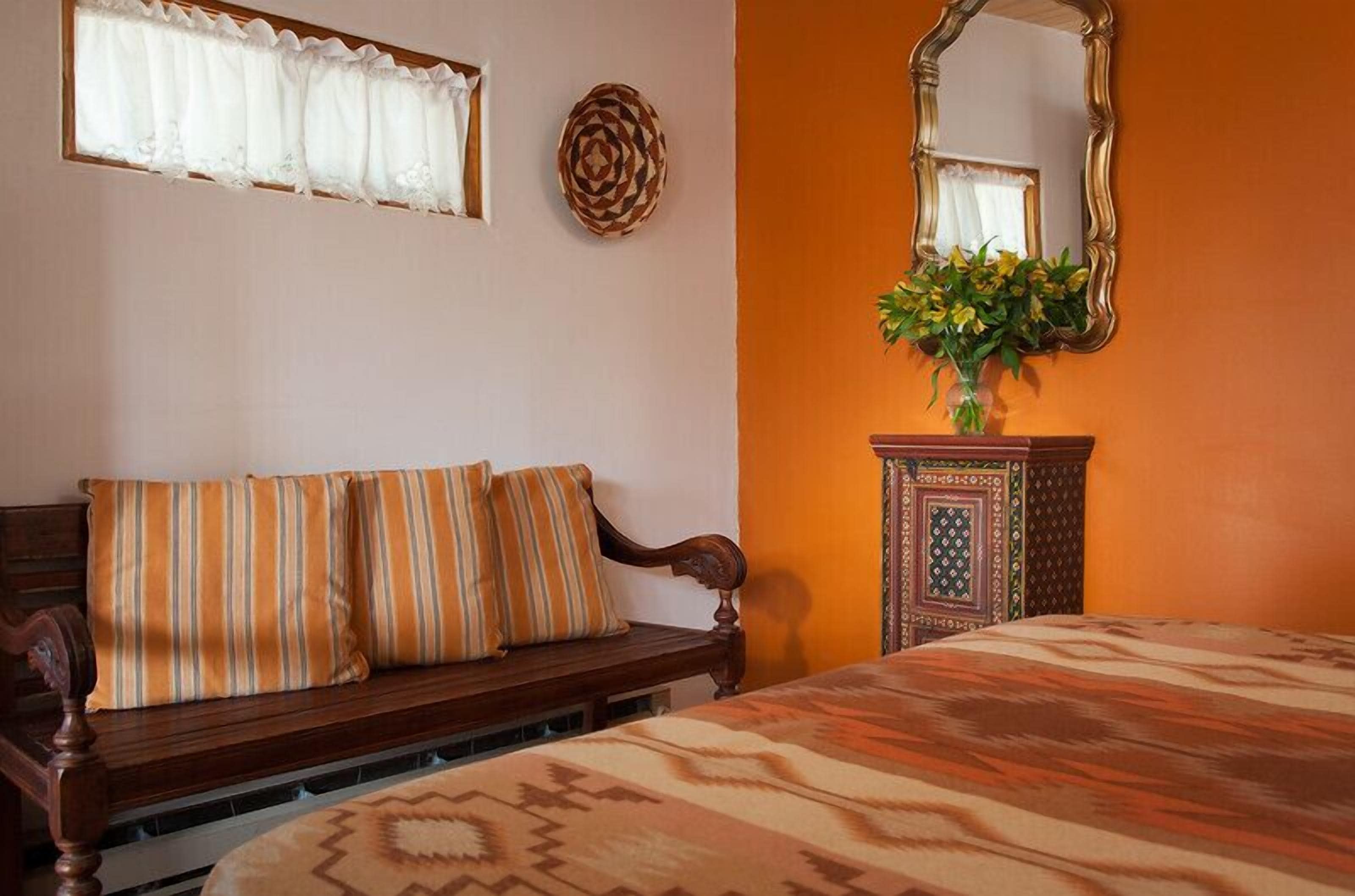 A bedroom with a wooden bench, a patterned bedspread, a mirror, and a vase with flowers.