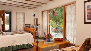 A cozy room with a bed, a wooden table with a vase of flowers, a chair, and a view of greenery through large windows. - Casa Escondida Bed & Breakfast (Chimayo)