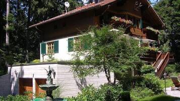 A traditional chalet with a sloped roof, a wooden staircase, and a small garden with a fountain.