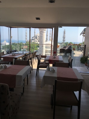 Un espace de restauration en plein air avec des tables et des chaises, de grandes fenêtres offrant une vue sur une mosquée et la mer.