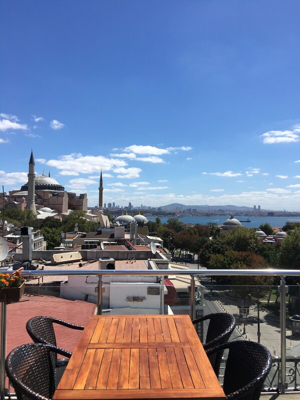 A terrace with a wooden table and wicker chairs, overlooking a cityscape with historic buildings and a river.