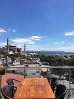 Una terraza con una mesa de madera y sillas de mimbre, con vistas a un paisaje urbano que incluye edificios históricos y un río.