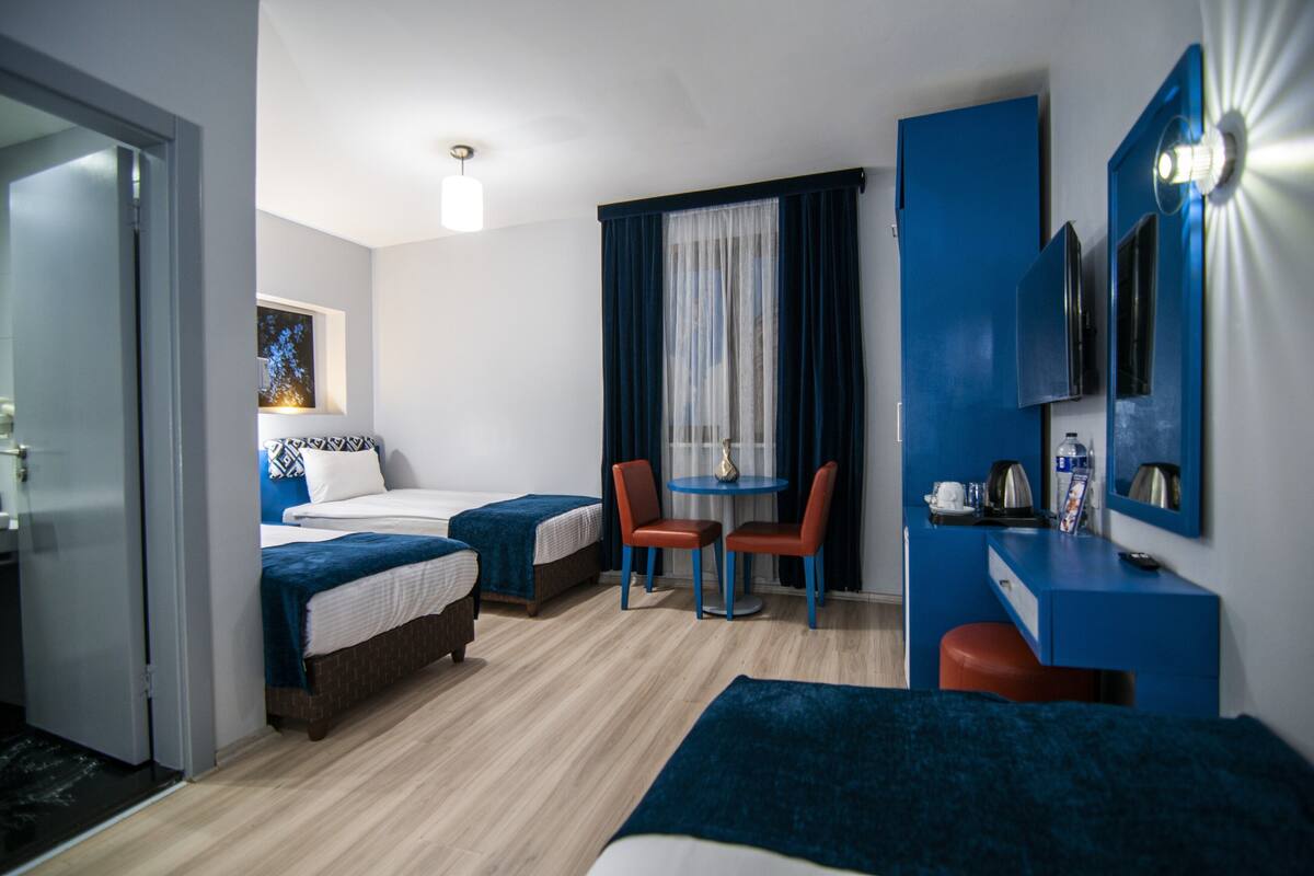 A hotel room with two beds, a small table with chairs, a desk with a chair, a TV, and a window with curtains.