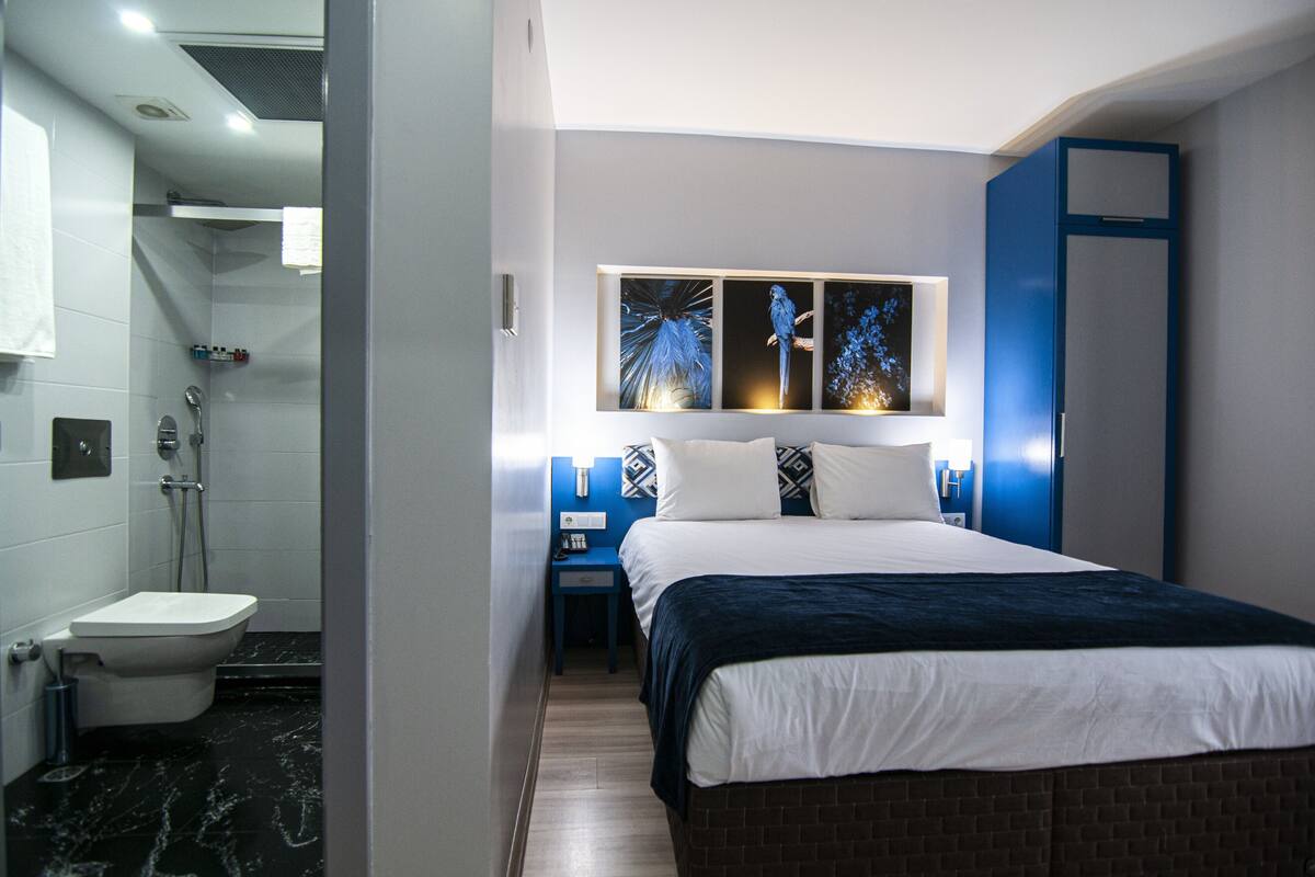 A modern hotel room with a bed, a nightstand, a bathroom with a toilet and shower, and a wall-mounted artwork.