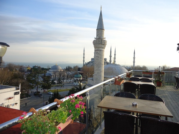 Une terrasse sur le toit offrant une vue sur une mosquée, notamment sur son dôme et ses minarets. On y trouve des tables et des chaises pour prendre ses repas, ainsi que des plantes en pot disposées sur la balustrade.