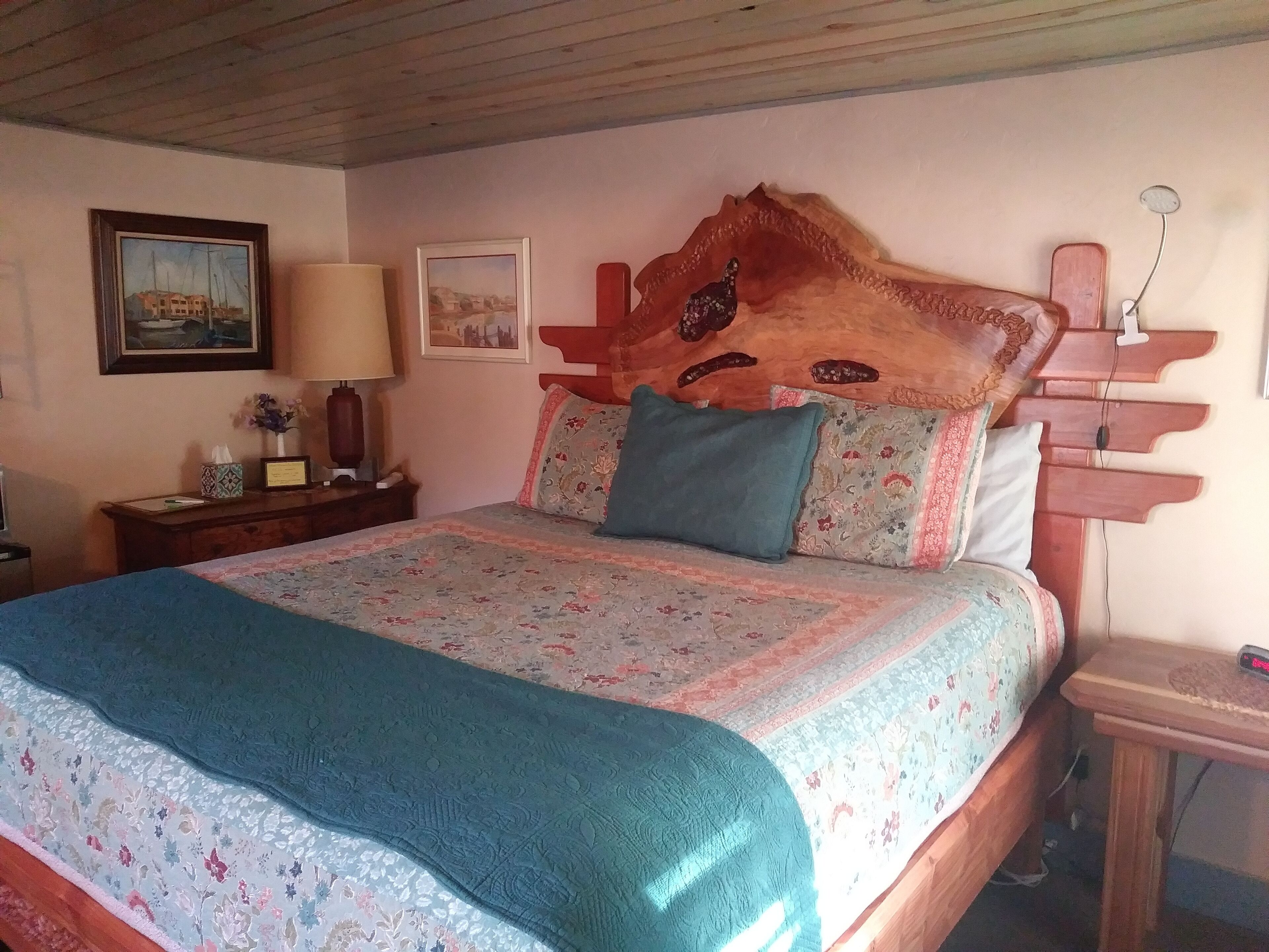 A bedroom with a wooden headboard, floral bedding, a bedside table with a lamp, and framed pictures on the wall.