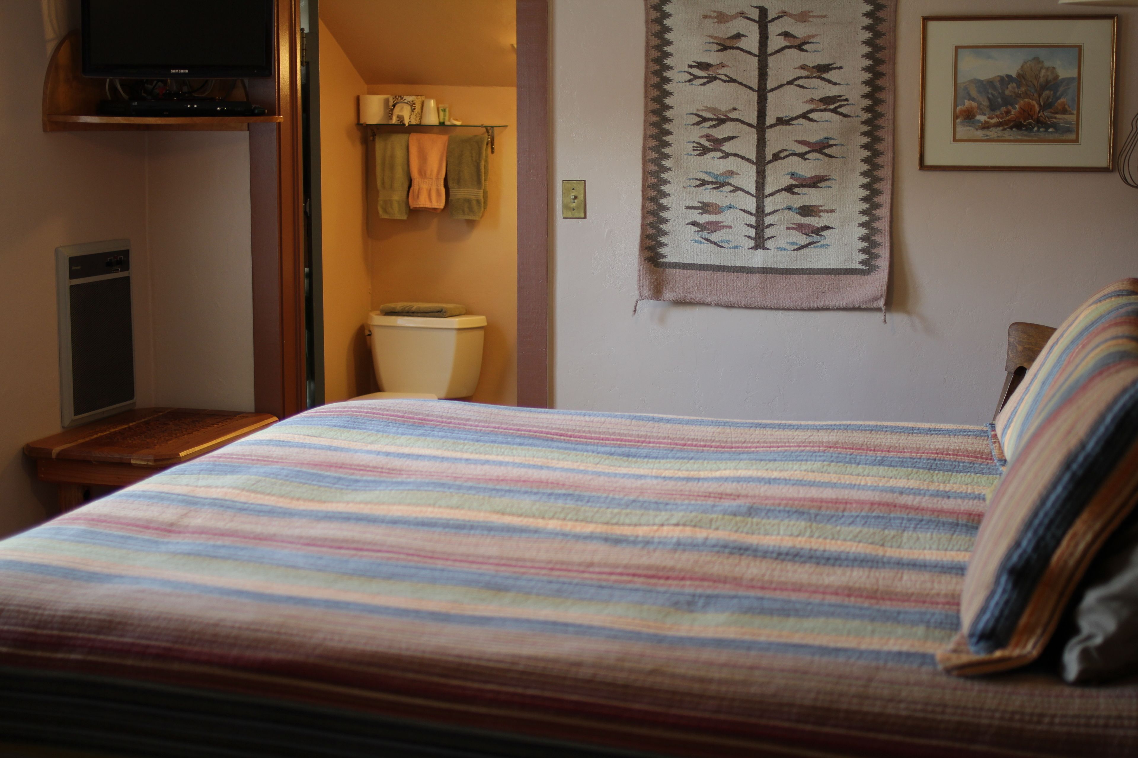 A bedroom with a bed, a television on a stand, a bathroom with a toilet, and a wall-mounted painting.