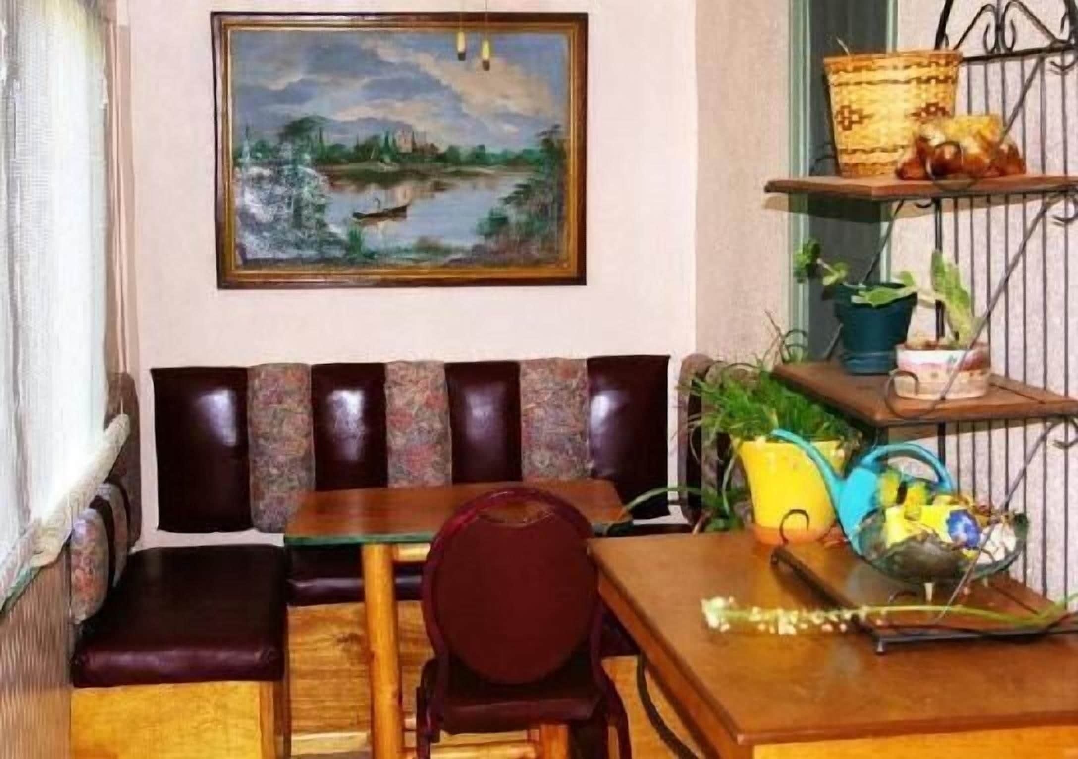 Un coin salon accueillant avec un banc rembourré, une table en bois et une chaise. Un tableau est accroché au mur, au-dessus du banc, et une étagère contient des plantes et des objets décoratifs.