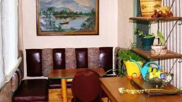 Un coin salon accueillant avec un banc rembourré, une table en bois et une chaise. Un tableau est accroché au mur, au-dessus du banc, et une étagère contient des plantes et des objets décoratifs.