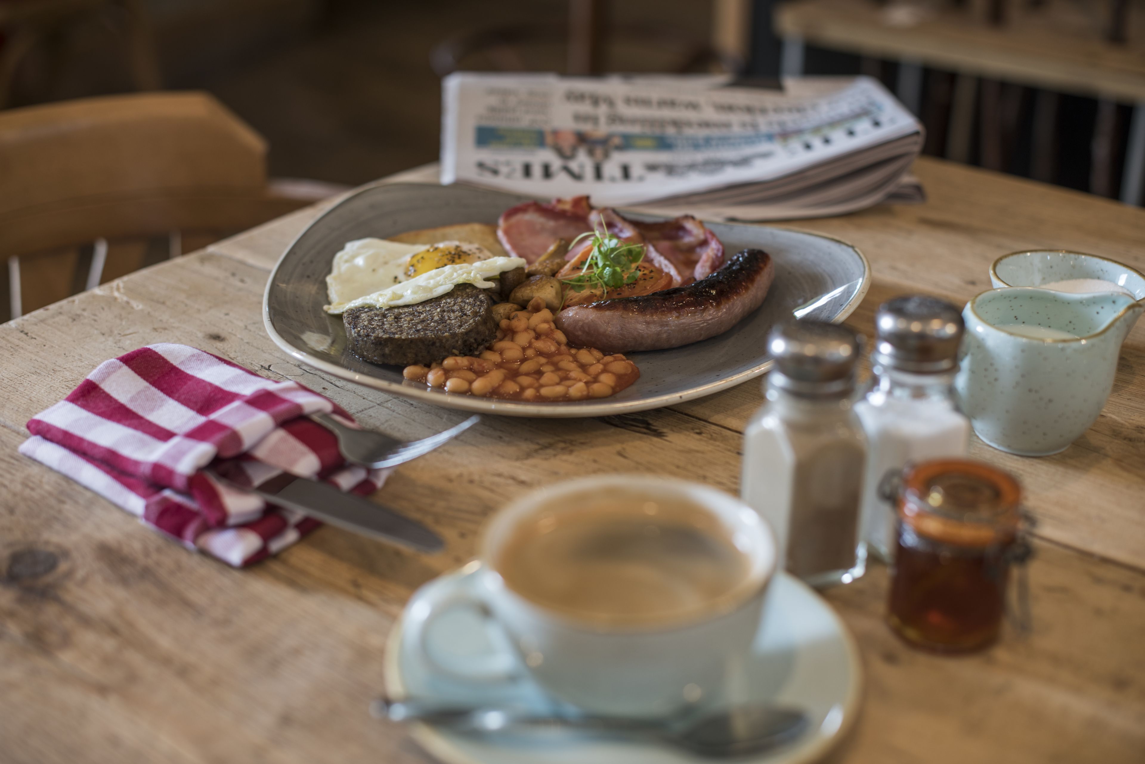A breakfast plate with eggs, beans, and bacon, a cup of coffee, salt and pepper shakers, and a newspaper on a wooden table.