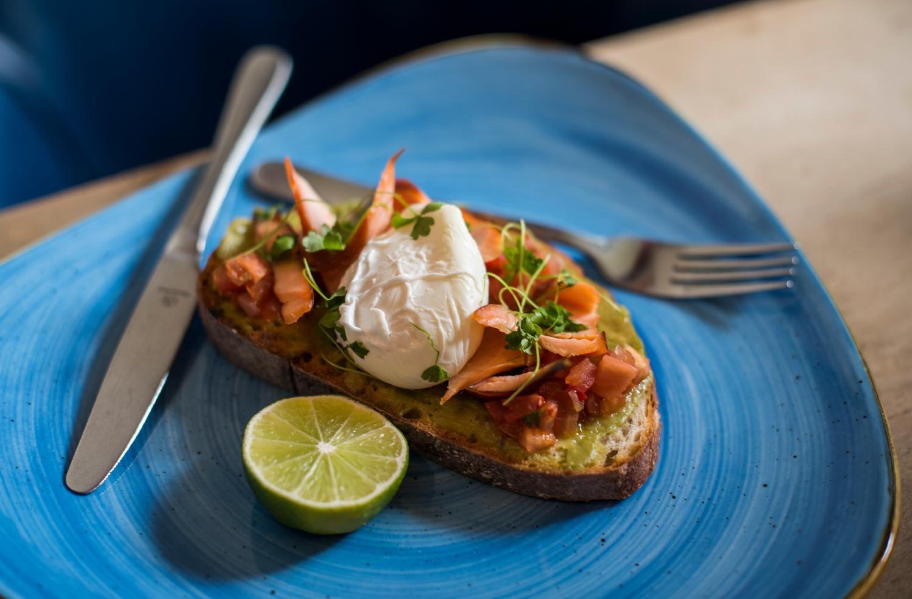 A plate with a slice of toast topped with salmon, a dollop of cream, and garnished with herbs, accompanied by a slice of lime.