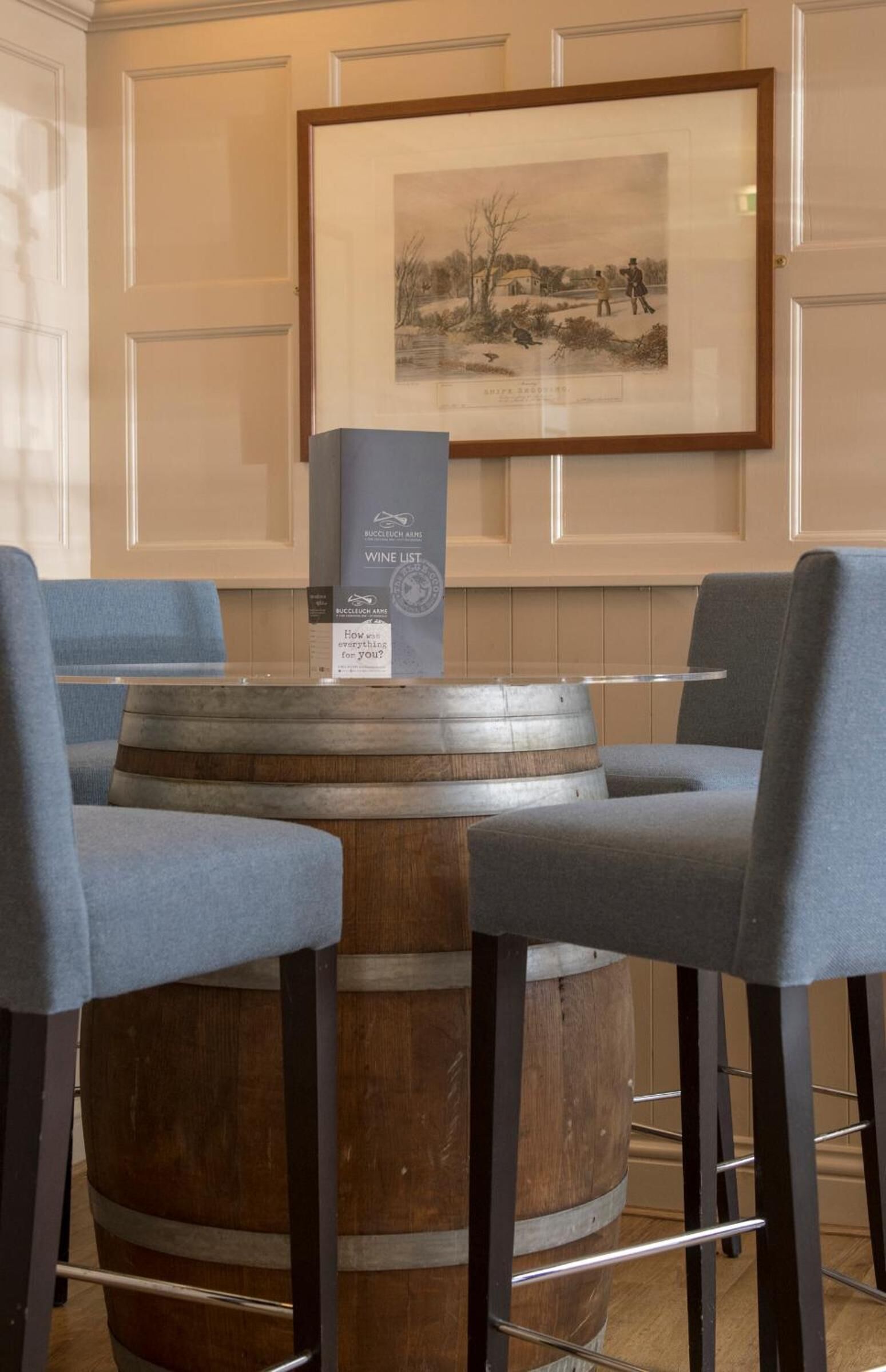 Un coin bar avec un tonneau en bois, deux chaises bleues, une œuvre d’art encadrée au mur et un présentoir pour la carte des vins.