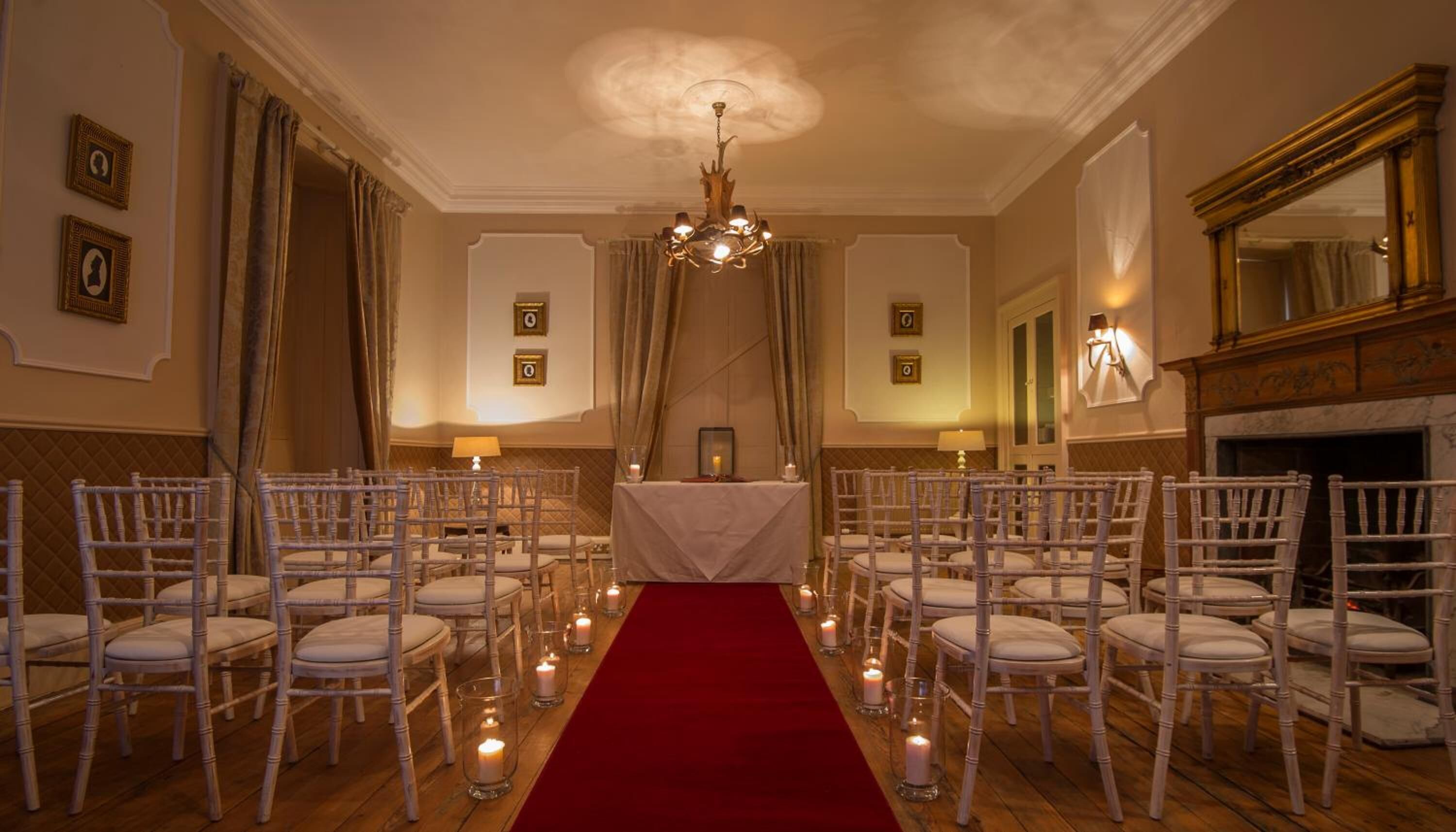 A formal room with a red carpet aisle, white chairs, a chandelier, framed pictures, and a fireplace.