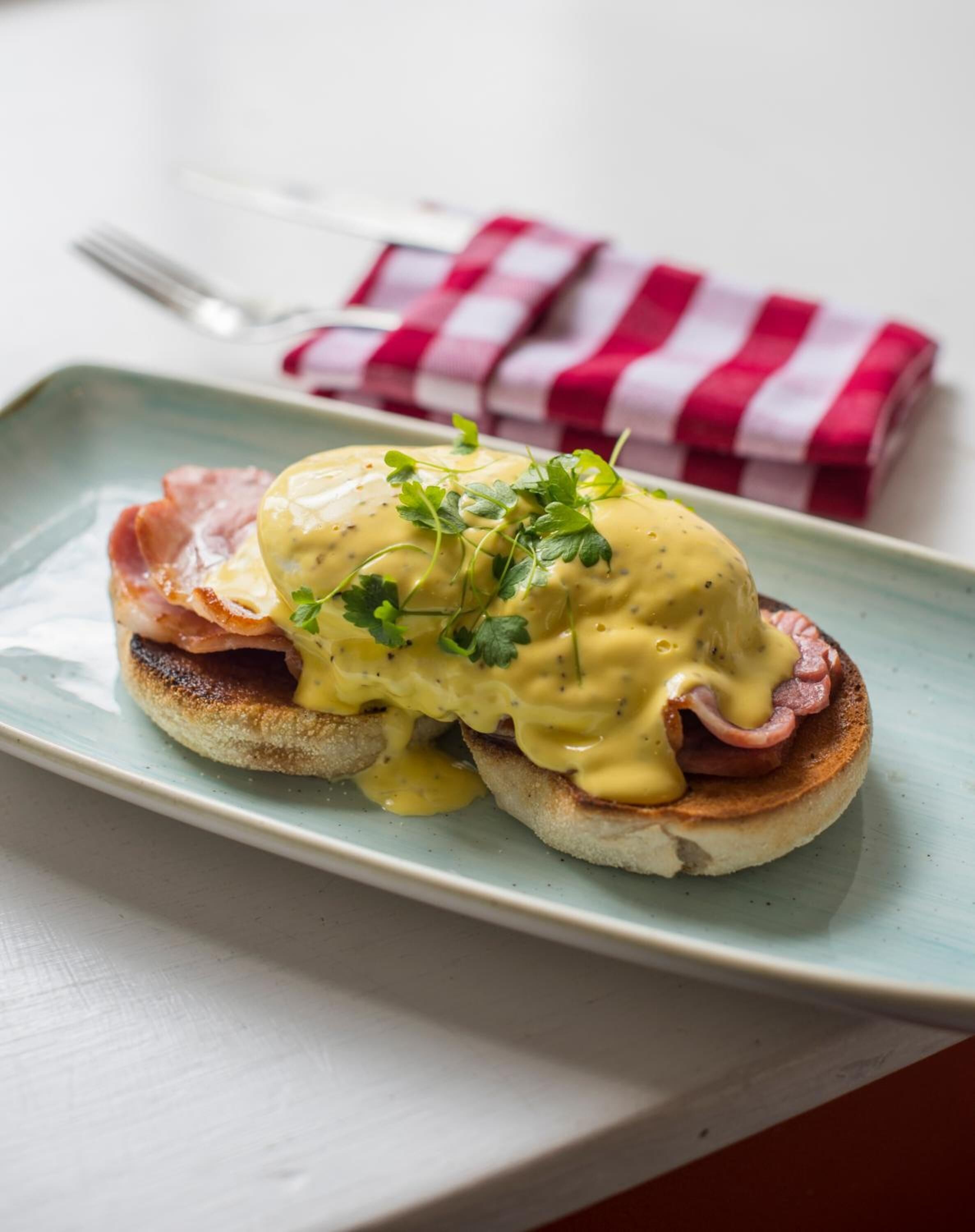 Une assiette d’œufs Bénédicte, accompagnés de jambon et de sauce hollandaise, décorée de persil, le tout présenté sur une assiette bleu clair.