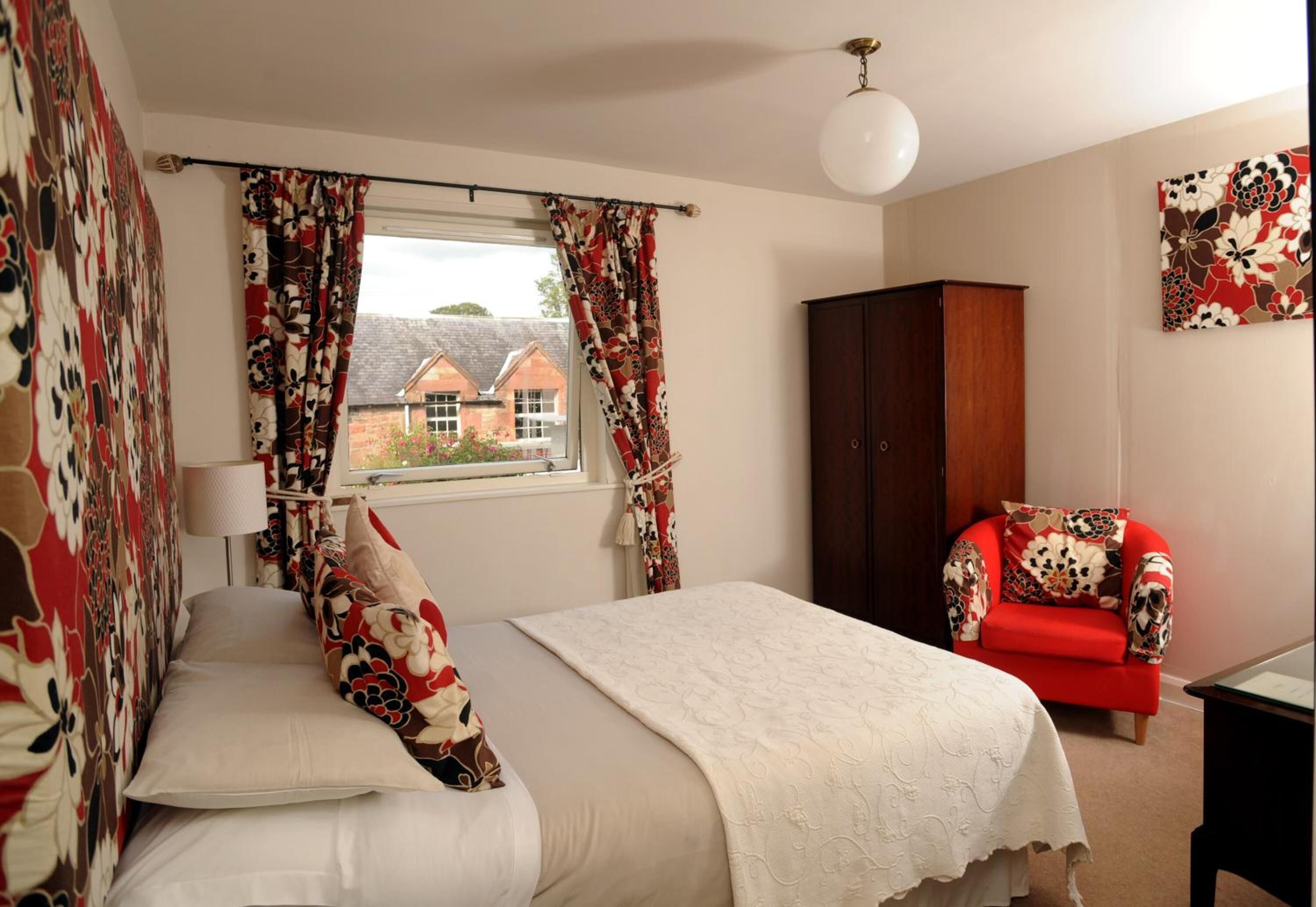 A bedroom with a bed, a window with curtains, a red chair, and a wooden wardrobe.