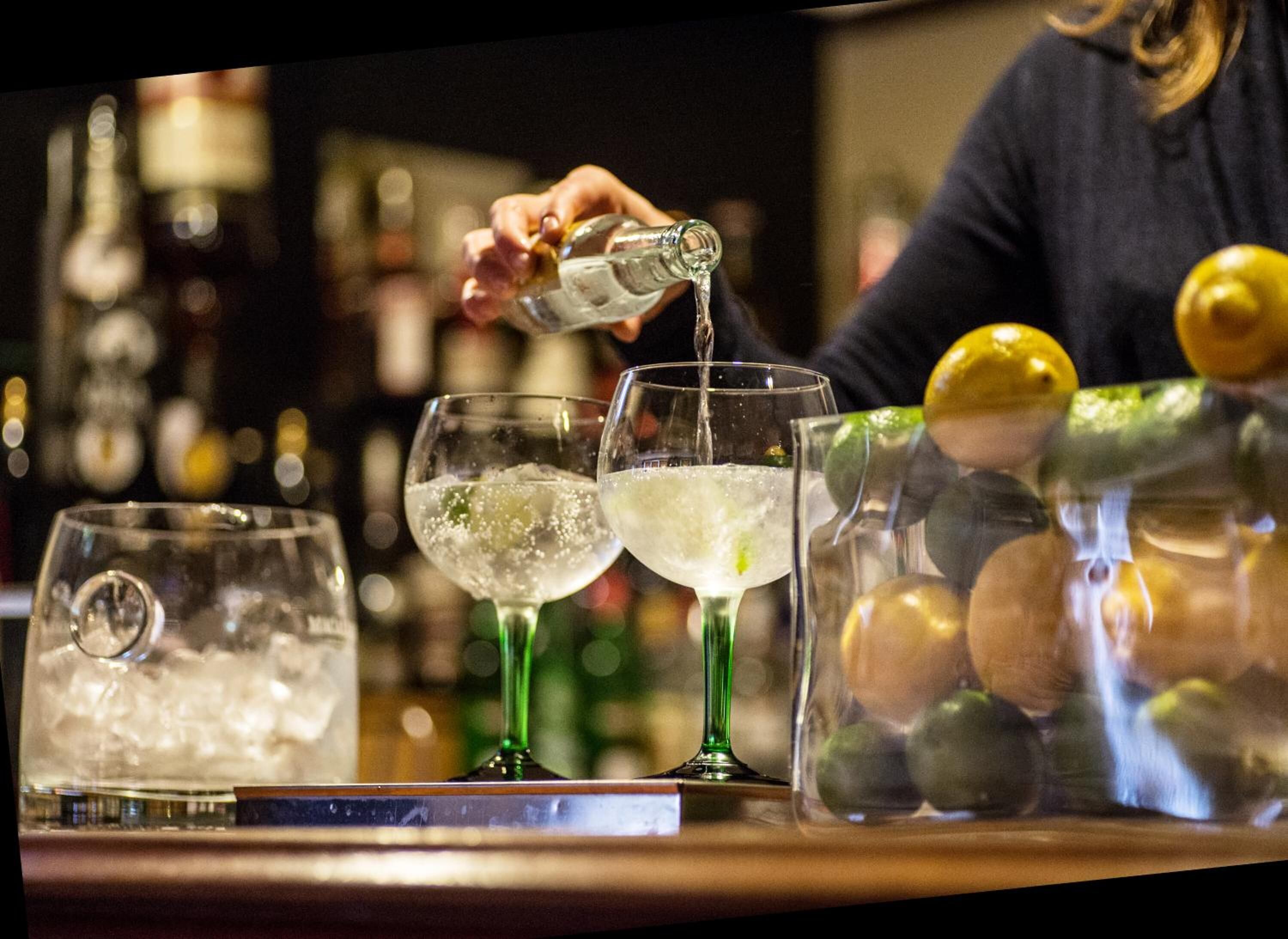 A bar setting with a bartender pouring a drink into a glass. There are ice cubes in the glasses, and a pitcher of lemons is visible.