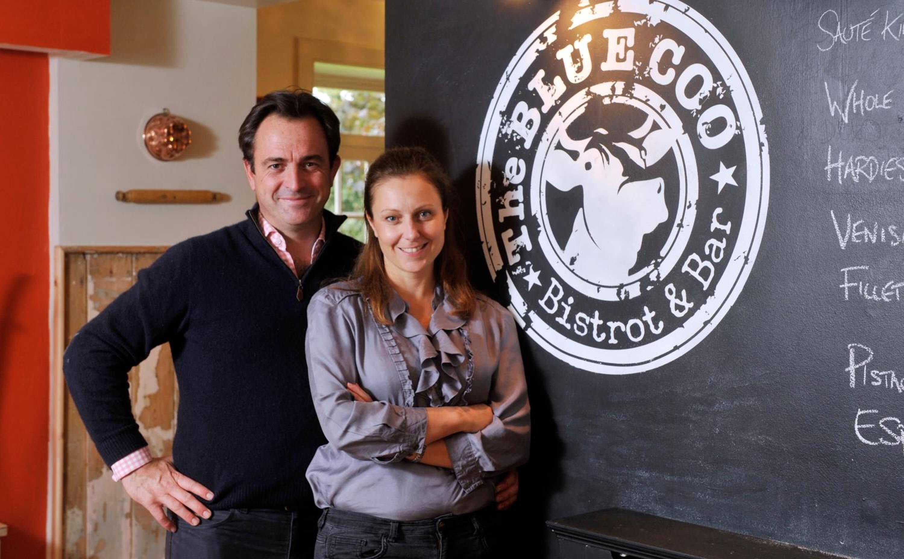Two people standing in front of a wall with a chalkboard listing items like "Whole Hardies" and "Venis."