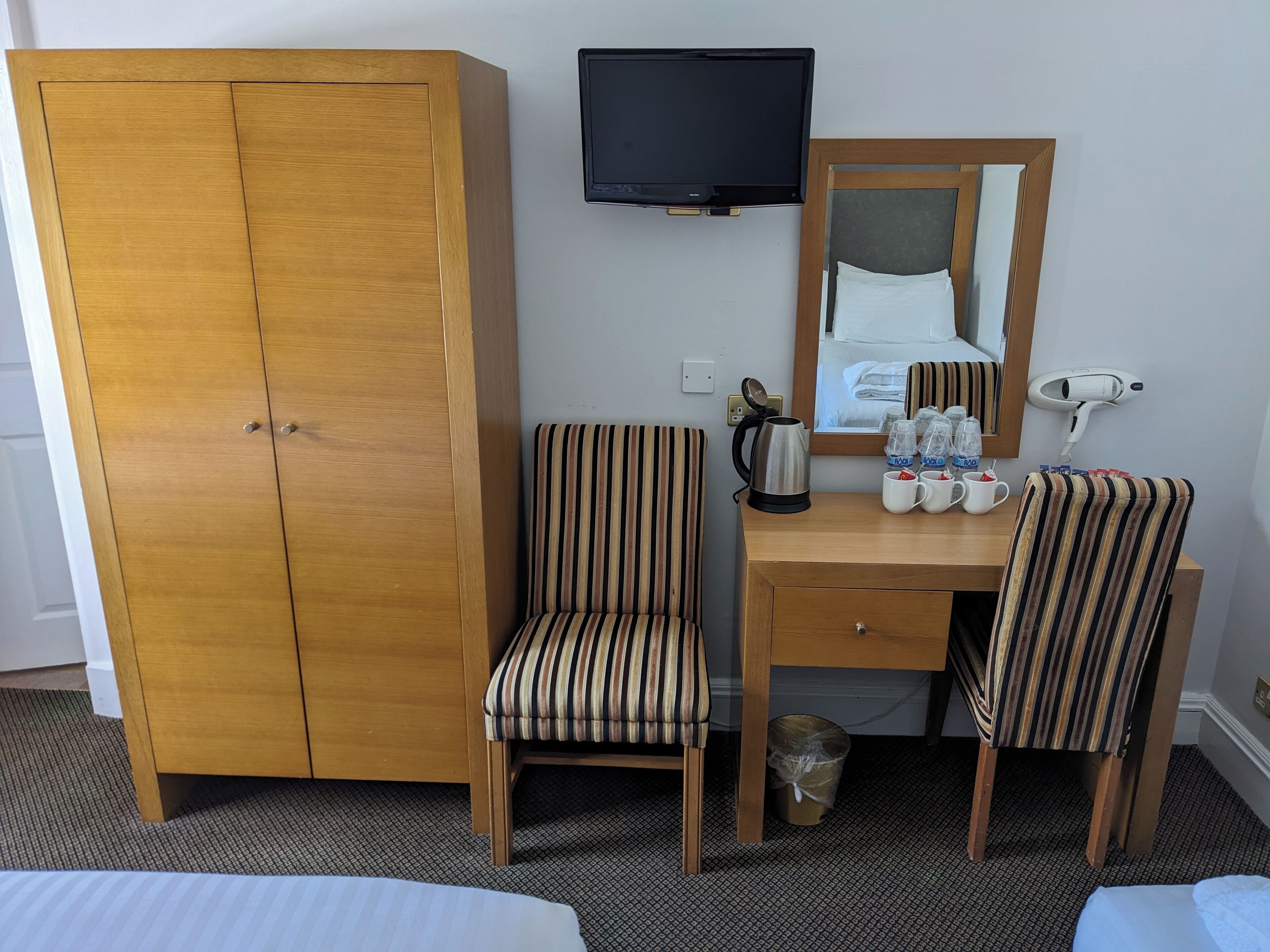 Een hotelkamer met een houten garderobe, een bureau met twee gestreepte stoelen, een televisie, een spiegel en een haardroger.