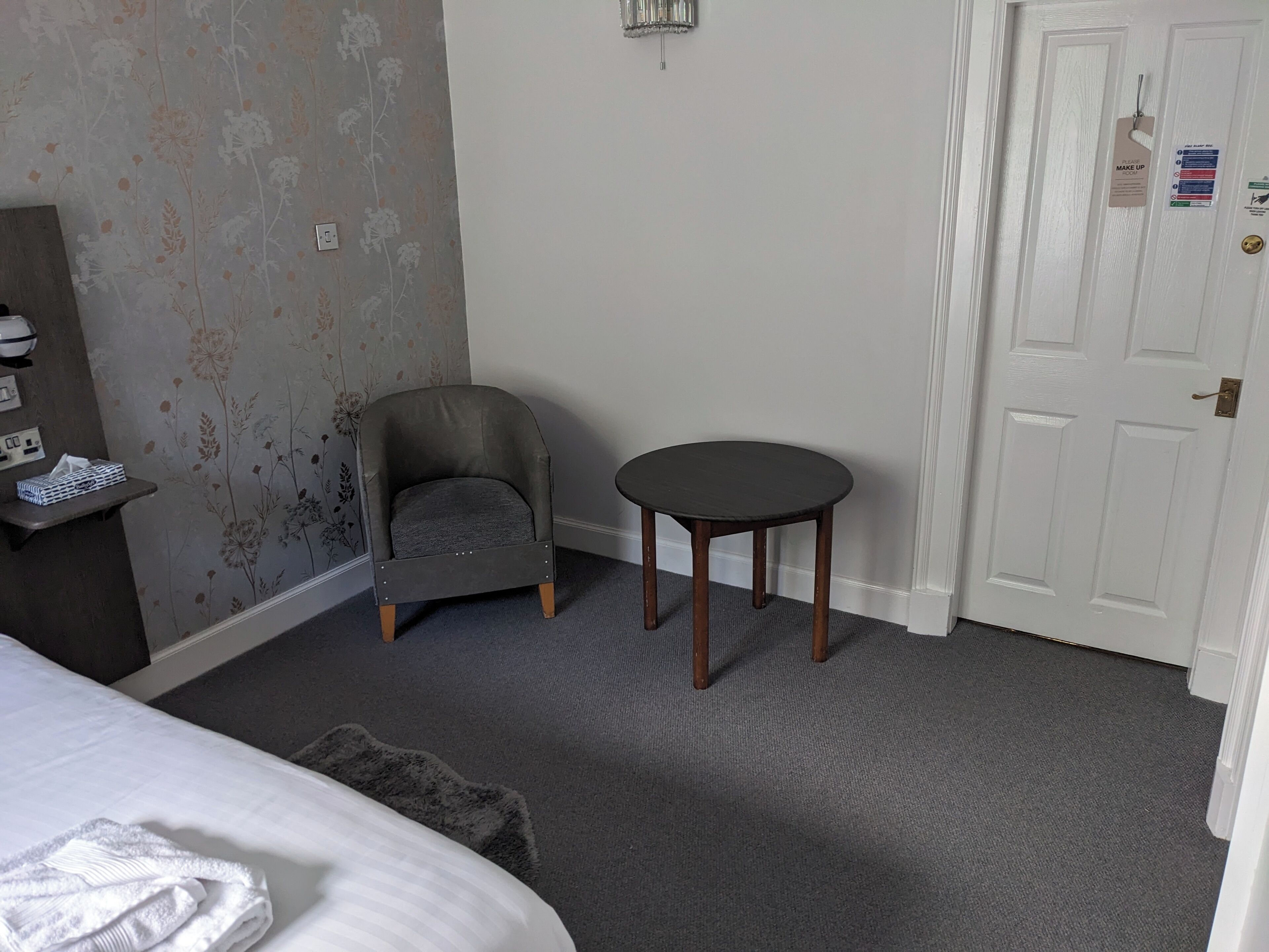 Une chambre d’hôtel avec un papier peint à motifs floraux gris, un fauteuil gris, une petite table ronde, une porte blanche avec une pancarte indiquant « MAQUILLAGE », et un lit recouvert d’une couverture blanche.