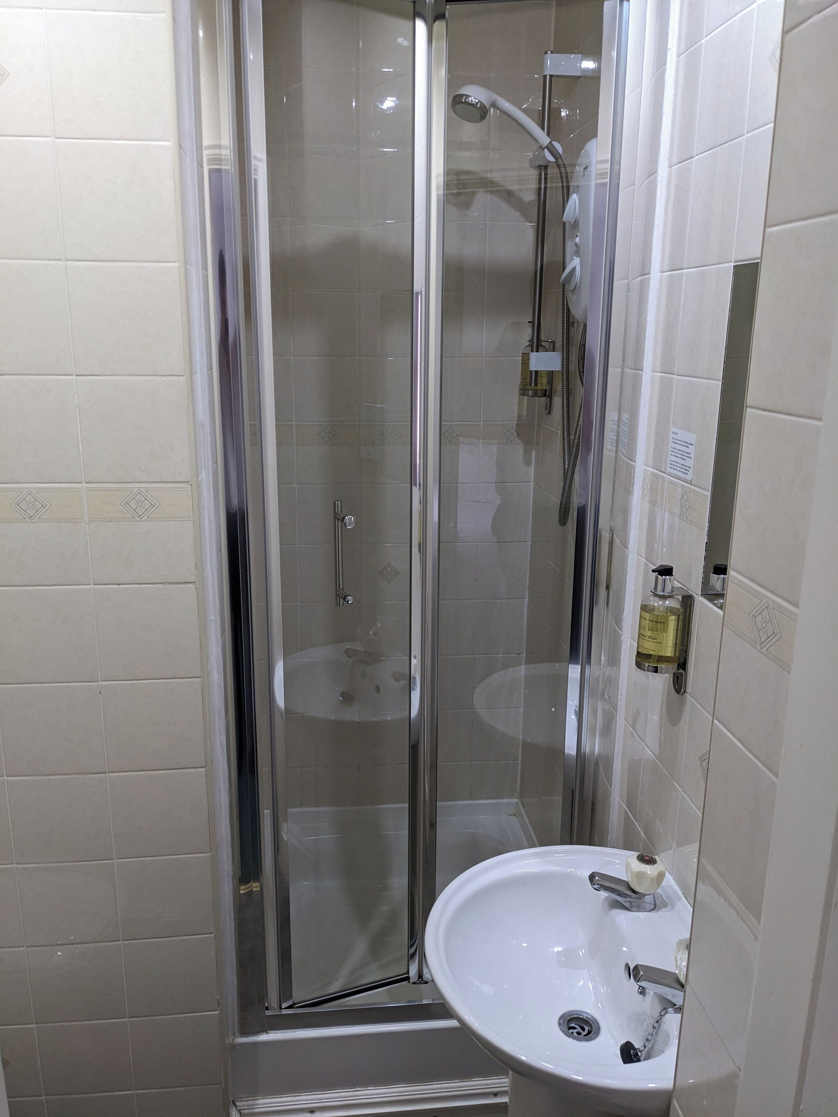 A glass-enclosed shower with a handheld showerhead and a wall-mounted faucet. Adjacent to the shower is a white sink with a single faucet.