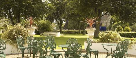 A courtyard with green metal chairs and tables, surrounded by lush greenery and a well-maintained lawn.