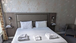 En pænt redt seng med hvide sengetøj og to puder, en grå sengegavl og en grå lænestol i et hotelværelse.