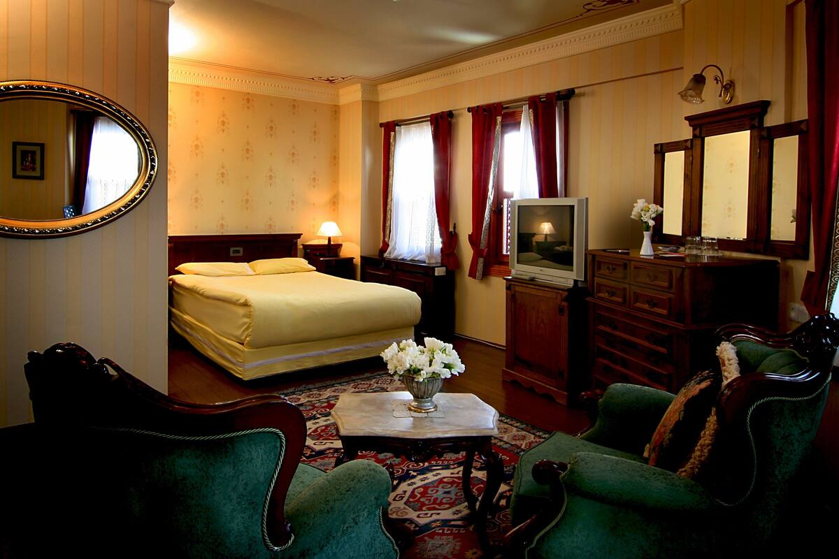 A hotel room with a bed, a television, a desk with a vase of flowers, and green upholstered chairs.