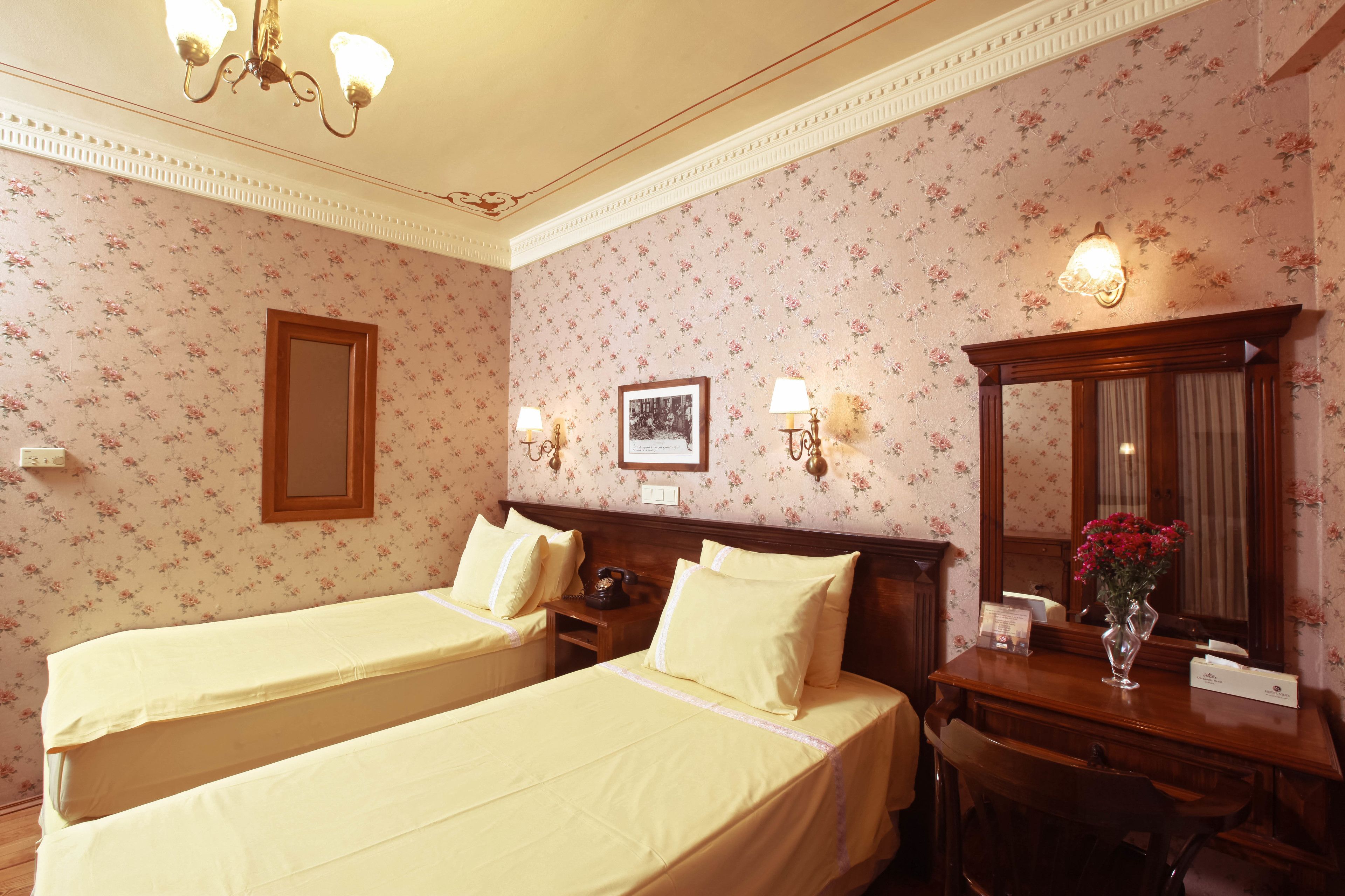 A hotel room with two beds, a wooden desk, a mirror, and floral wallpaper.