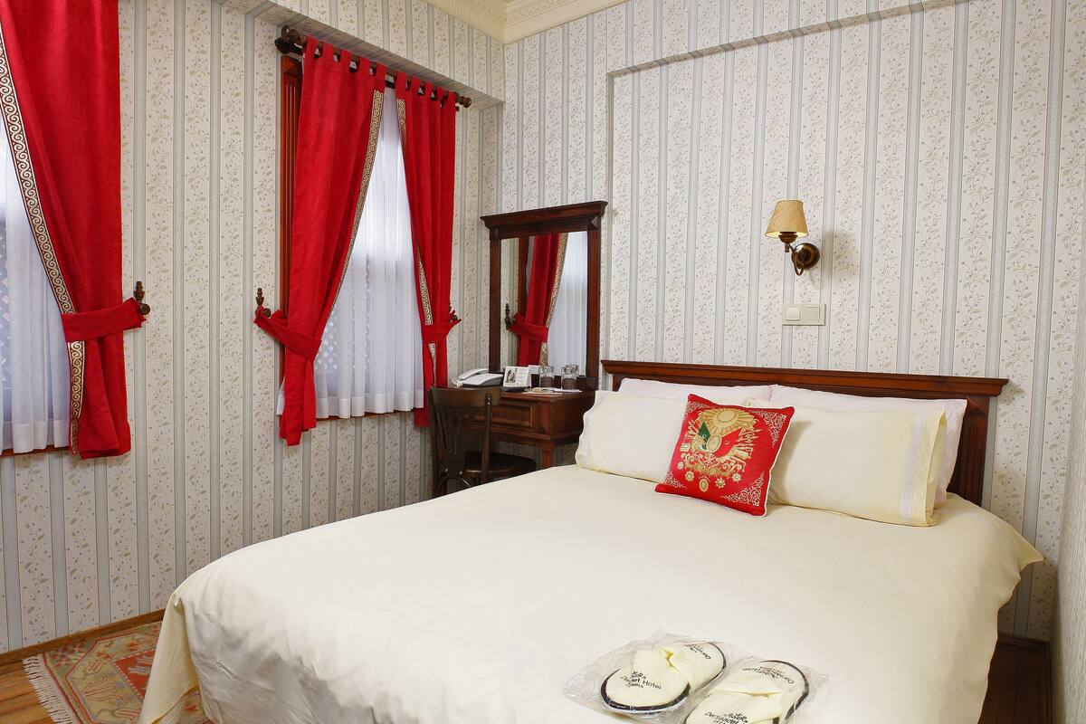 A hotel room with a large bed, red curtains, a wooden headboard, a bedside lamp, and a mirror.