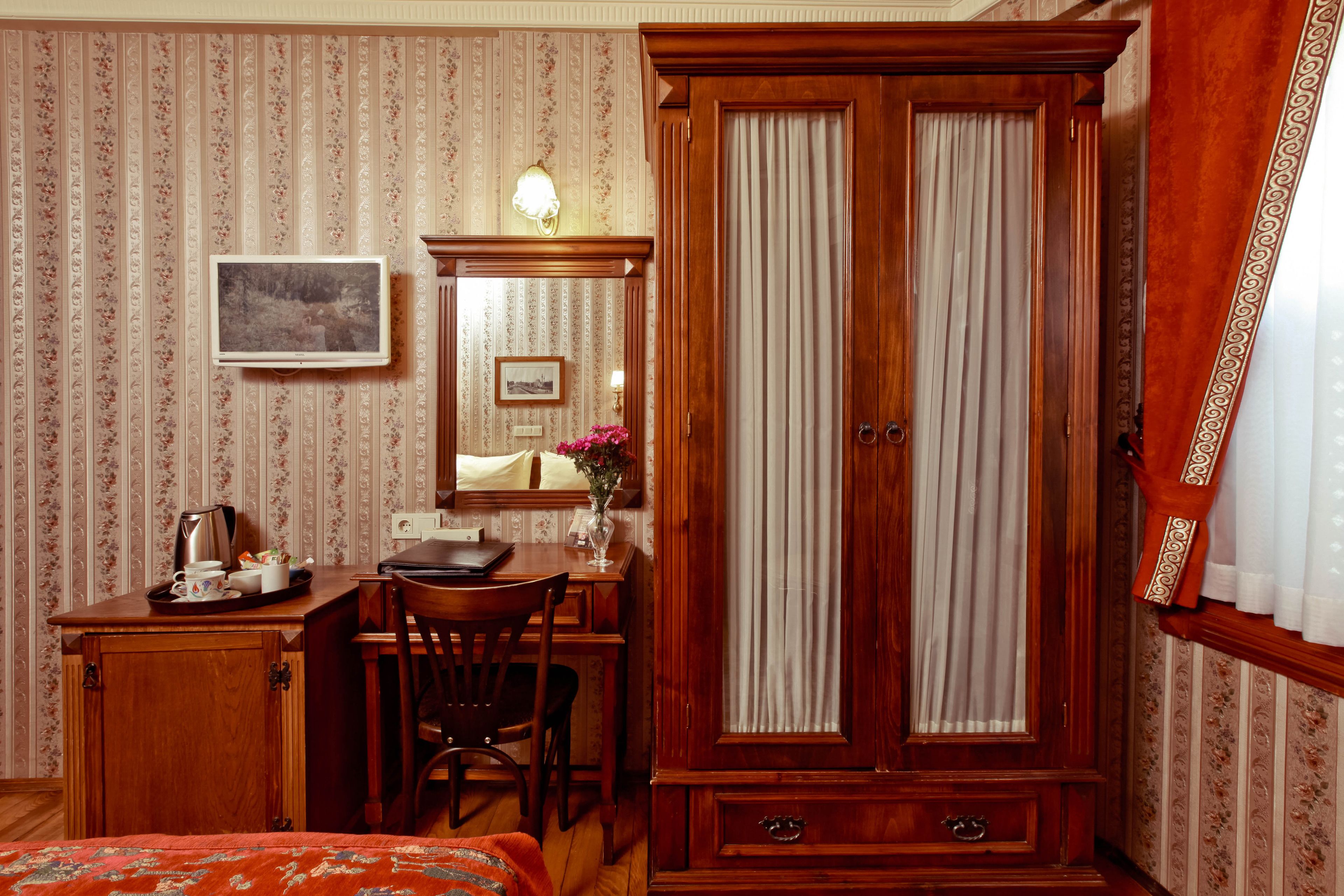 A room with a wooden wardrobe, a desk with a chair, a bed, and a wall-mounted picture.