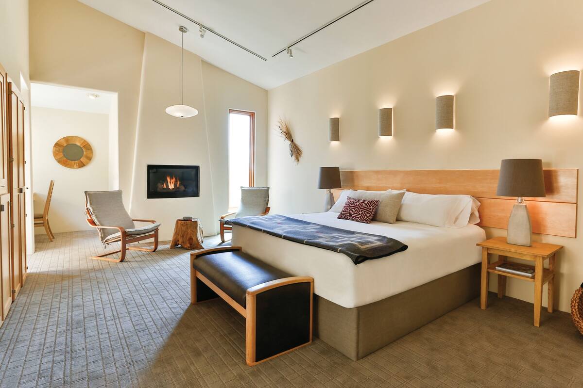 A modern hotel room with a large bed, a bench, a fireplace, and a view of the city.