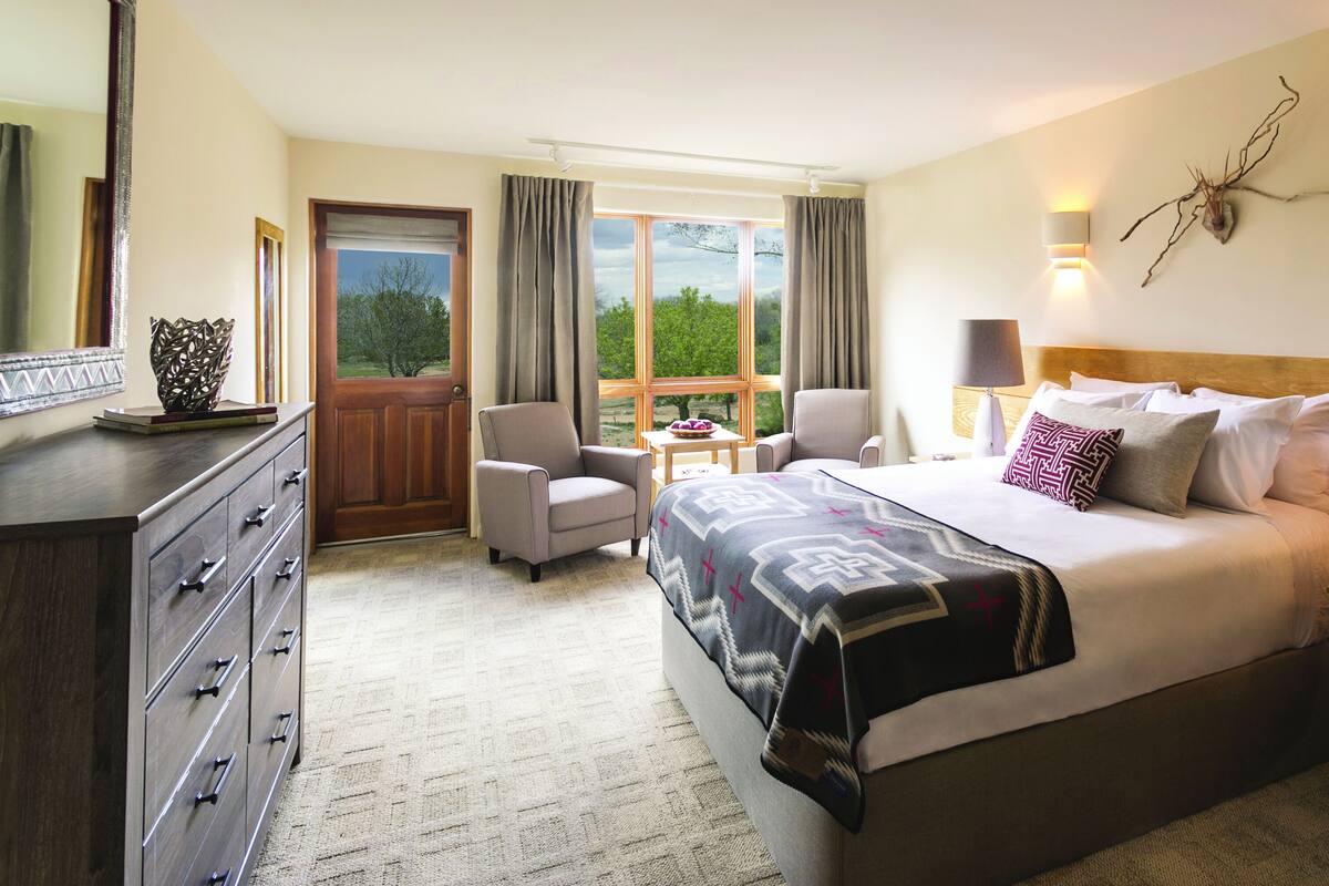 A hotel room with a large bed, two chairs, a wooden dresser, a table with a bowl, and a window with curtains.