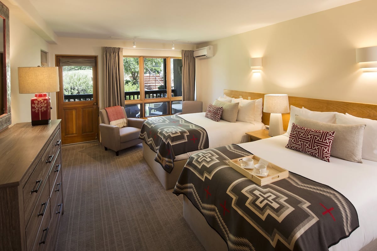 A hotel room with two beds, a desk, a chair, and a large window with a view of greenery.