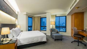 A hotel room with a bed, desk, chair, and large windows offering a city view.