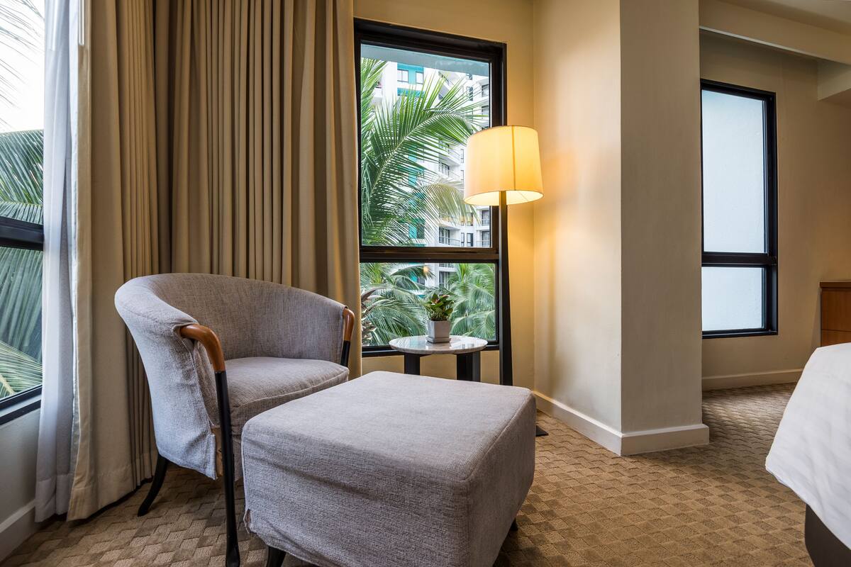 Ein Hotelzimmer mit großem Fenster, einer Lampe, einem Sessel, einem kleinen Tisch mit Pflanze und einem Bett.