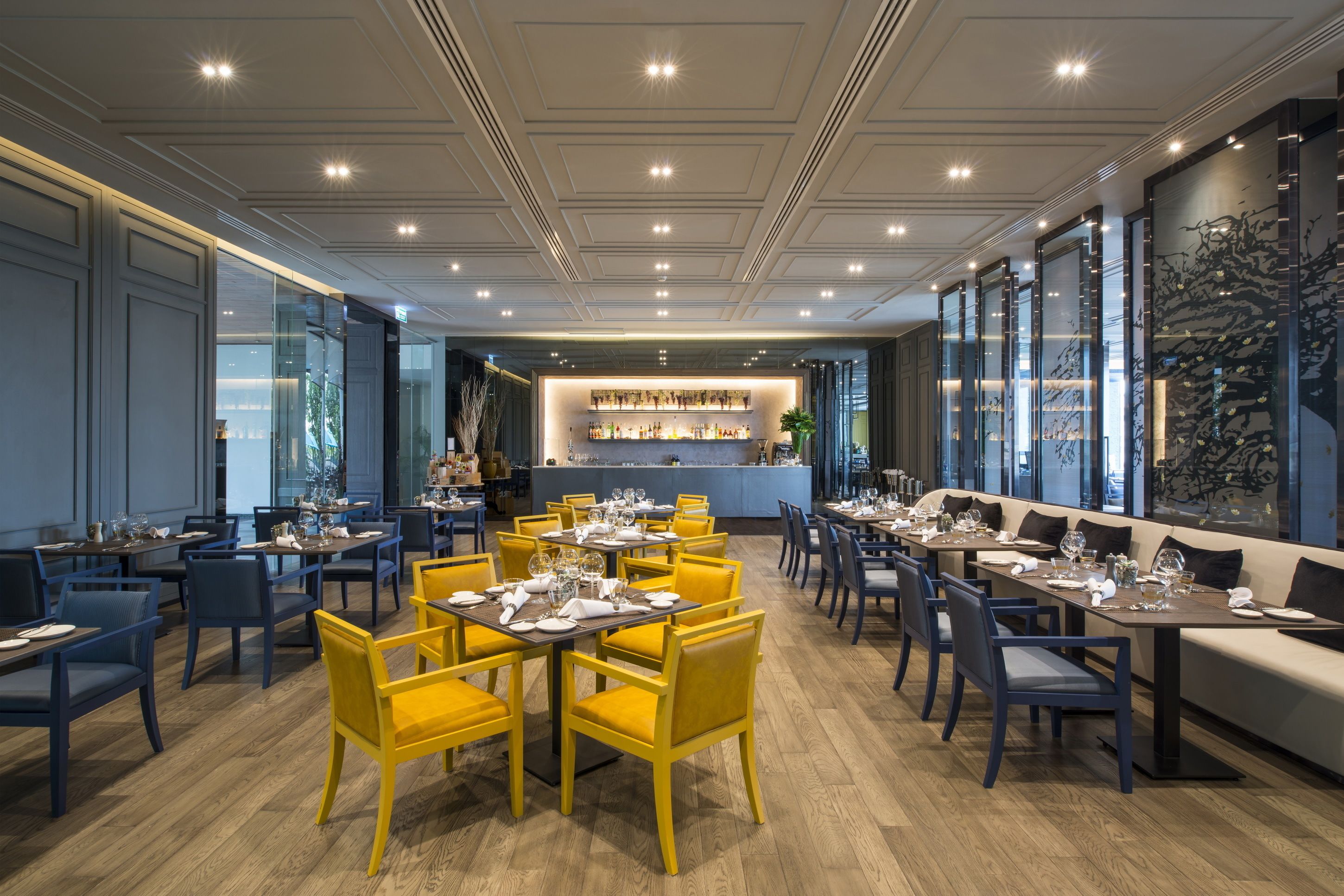 A modern restaurant with yellow and blue chairs, a bar area, and large windows.