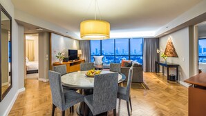 Una habitación de hotel moderna con zona de comedor, televisión y un amplio ventanal con vistas a la ciudad.