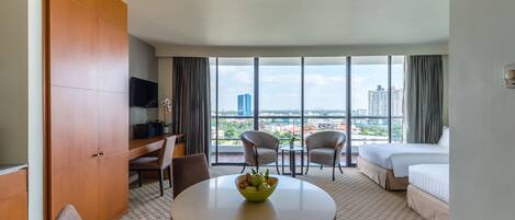 A hotel room with a large window, a bed, a desk with a TV, two chairs, and a round table with a fruit bowl.