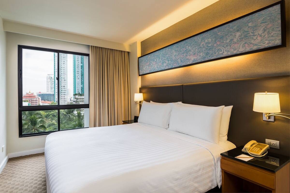 Ein Hotelzimmer mit einem großen Bett, einem Nachttisch mit Telefon, einer Wandlampe und einem Fenster mit Blick auf hohe Gebäude und Palmen.