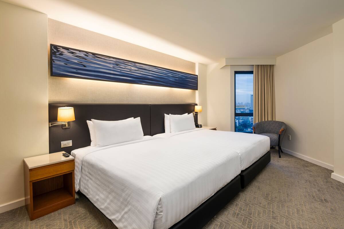 Ein Hotelzimmer mit einem großen Bett, zwei Nachttischlampen, einem Nachttisch, einem Sessel und Blick auf die Stadt durch ein Fenster.